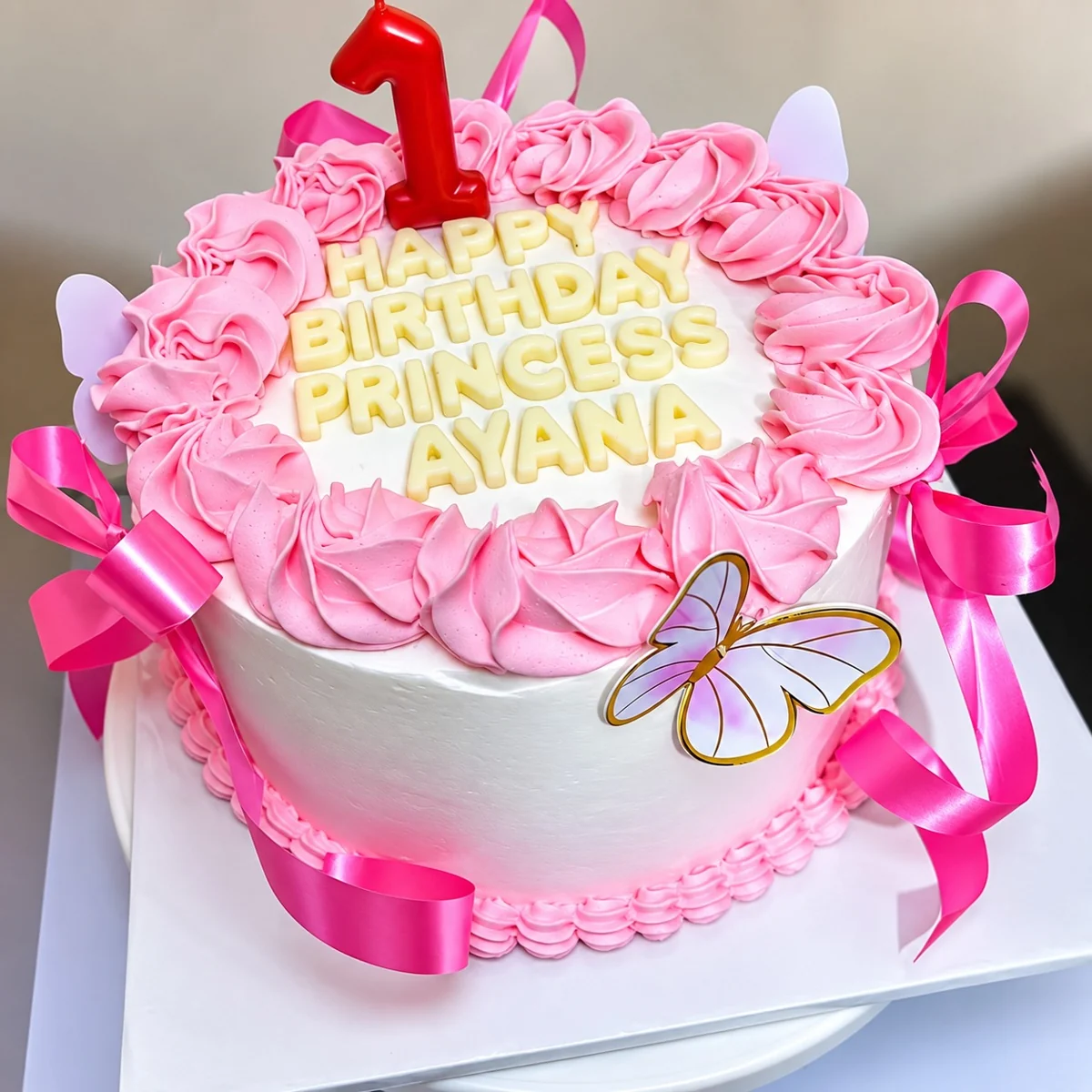 Round cake with pink piping, ribbons, butterflies and a number one topper by Moh Bakes & Treats