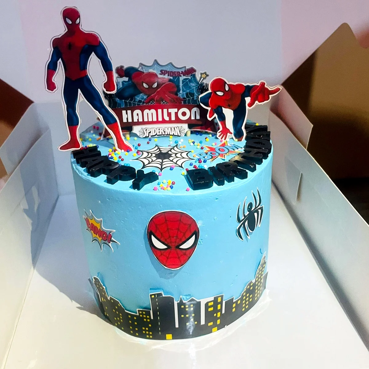 Round superhero cake with Spiderman toppers and a city skyline by Moh Bakes & Treats